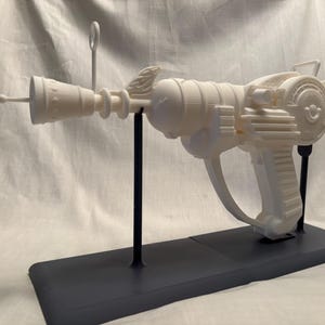 May include: A white ray gun prop, possibly 3D-printed, displayed on a black stand. The futuristic weapon has a cylindrical barrel, detailed body, and a trigger. The design suggests a science fiction or retro aesthetic.