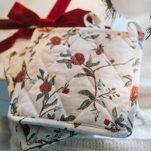 May include: A white quilted pot holder with a floral pattern featuring red berries and green leaves. The pot holder has a red ribbon tied around it.