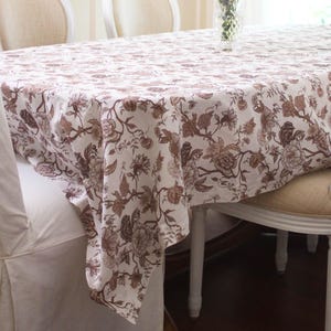 May include: A dining table set with a floral patterned tablecloth in shades of brown and white. The table is surrounded by white chairs with beige upholstered seats. A clear glass vase with blue flowers sits in the center of the table.