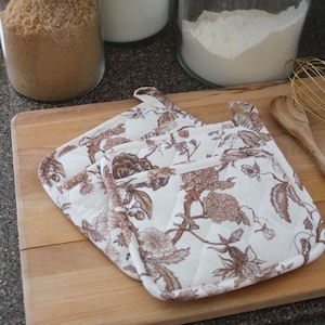May include: Two white and brown floral patterned oven mitts rest on a wooden cutting board. Glass jars filled with brown sugar, white sugar, and flour are in the background. A whisk and wooden spoon are also visible.