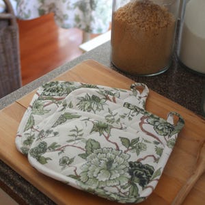 May include: Two white and green floral print pot holders with a quilted design rest on a wooden cutting board. A glass jar filled with brown sugar and a second jar are in the background.