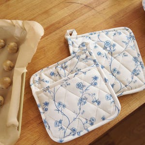 May include: White quilted oven mitts and pot holders with a blue floral pattern. A baking sheet with cookie dough balls is on a wooden surface. Kitchen and baking supplies.