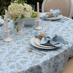 May include: A table setting with a light blue floral tablecloth, white plates, and blue napkins. White candles in ceramic holders and a white vase with flowers are also present. Silverware and wine glasses complete the elegant outdoor dining scene.