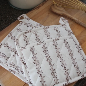 May include: Two white quilted pot holders with a floral pattern in brown. The pot holders are square with a loop for hanging. They are placed on a wooden cutting board, with a whisk and wooden spoon in the background.