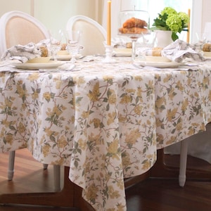 May include: A dining table set for a meal, featuring a white tablecloth with a yellow floral pattern. The table is set with plates, napkins, and glassware. A lit candle and a glass dome with pastries are also present.