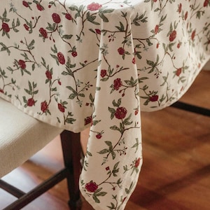 May include: A white tablecloth with a floral pattern featuring red flowers and green leaves. The tablecloth is draped over a table and chair.