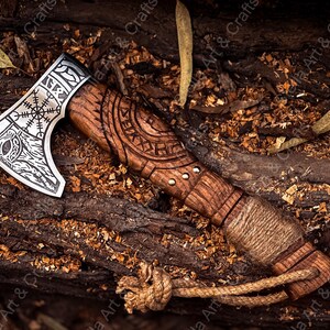 K&ouml;nnte beinhalten: Eine silberne Axt mit einem Holzstiel. Der Axtkopf hat ein detailliertes Design mit einem Kompass und Runen. Der Stiel ist mit braunem Leder umwickelt und hat ein Seil am Ende.