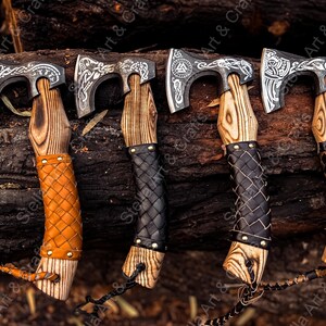 K&ouml;nnte beinhalten: Vier handgeschmiedete &Auml;xte mit aufwendigen Designs auf den Klingen und lederumwickelten Stielen. Die &Auml;xte werden auf einem Hintergrund aus Holzscheiten pr&auml;sentiert.