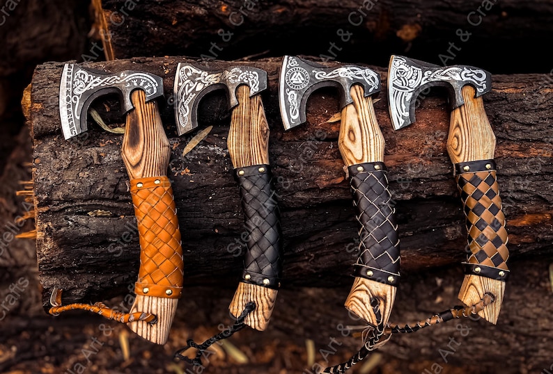 K&ouml;nnte beinhalten: Vier handgeschmiedete &Auml;xte mit aufwendigen Designs auf den Klingen und lederumwickelten Stielen. Die &Auml;xte werden auf einem Hintergrund aus Holzscheiten pr&auml;sentiert.