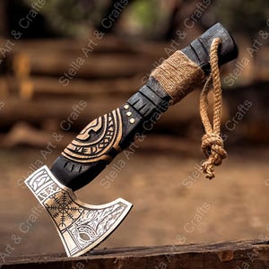 K&ouml;nnte beinhalten: Ein handgeschnitzter Axtkopf aus Holz mit einem schwarzen Stiel und einer silbernen Klinge. Die Klinge ist mit einem Wikinger-Knotenmuster und einem Wolfskopf verziert. Der Stiel ist mit braunem Garn umwickelt und hat eine Lederschlaufe.