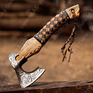 K&ouml;nnte beinhalten: Eine handgeschmiedete Axt mit einem Holzstiel, der mit Leder umwickelt ist. Die Axtkopf hat ein detailliertes Design und der Stiel ist mit einem Lederriemen befestigt. Die Axt liegt auf einer Holzoberfl&auml;che.