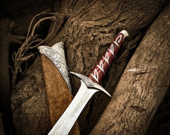 May include: A silver-toned metal sword with a red and silver patterned handle and a matching scabbard. The sword features an ornate crossguard with intricate engravings and a blade with etched details along the center and near the hilt.