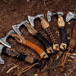 K&ouml;nnte beinhalten: Sechs handgeschmiedete Wikinger-&Auml;xte mit aufwendigen Designs und lederumwickelten Griffen. Die &Auml;xte haben eine schwarze und silberne Oberfl&auml;che und werden auf einem braunen Hintergrund pr&auml;sentiert.