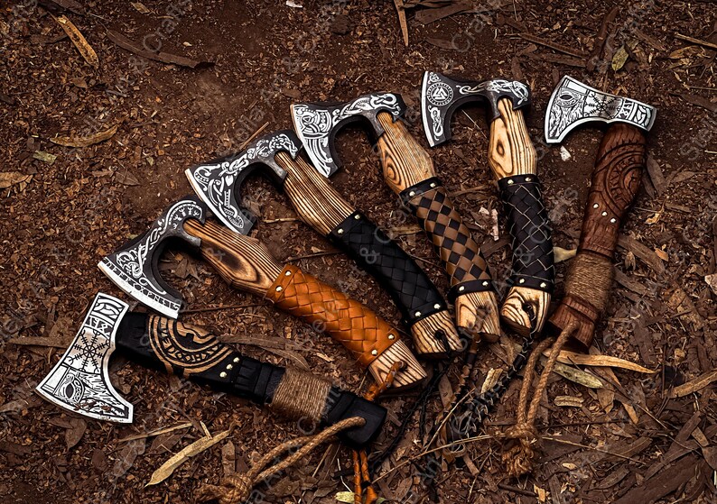 K&ouml;nnte beinhalten: Sechs handgeschmiedete Wikinger-&Auml;xte mit aufwendigen Designs und lederumwickelten Griffen. Die &Auml;xte haben eine schwarze und silberne Oberfl&auml;che und werden auf einem braunen Hintergrund pr&auml;sentiert.