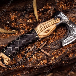 K&ouml;nnte beinhalten: Eine schwarze Axt mit einem Holzstiel und einer Lederummantelung. Der Axtkopf hat ein dekoratives Design mit einem Symbol eines Dreiecks mit einem Kreis darin. Die Axt liegt auf einem Bett aus Holzsp&auml;nen.