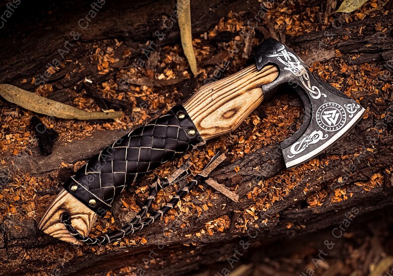 K&ouml;nnte beinhalten: Eine schwarze Axt mit einem Holzstiel und einer Lederummantelung. Der Axtkopf hat ein dekoratives Design mit einem Symbol eines Dreiecks mit einem Kreis darin. Die Axt liegt auf einem Bett aus Holzsp&auml;nen.