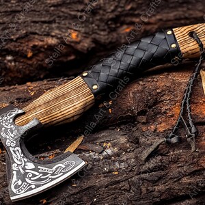 K&ouml;nnte beinhalten: Eine handgeschmiedete Axt mit einem Holzstiel und einer Lederummantelung. Der Axtkopf weist ein detailliertes, kunstvolles Design auf. Die Axt liegt auf einem Holzstapel.