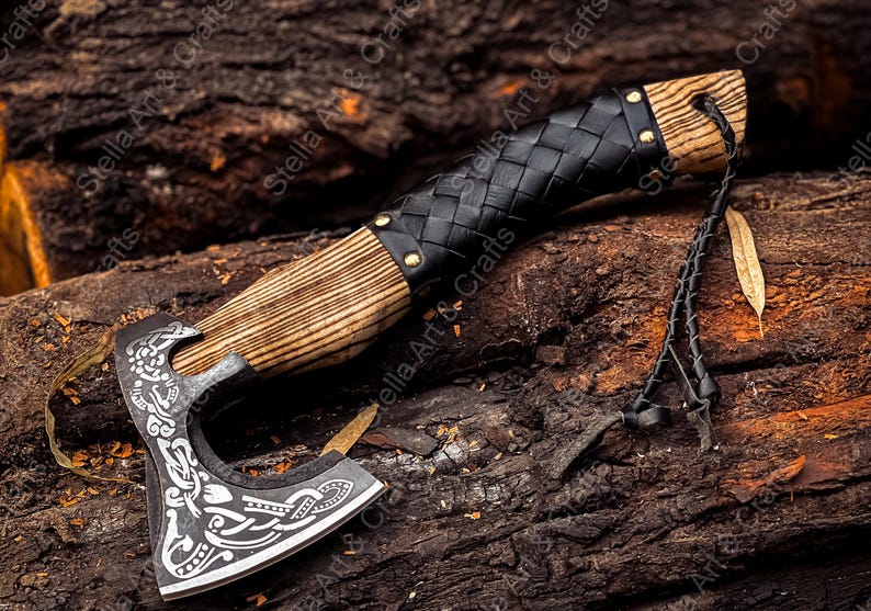K&ouml;nnte beinhalten: Eine handgeschmiedete Axt mit einem Holzstiel und einer Lederummantelung. Der Axtkopf weist ein detailliertes, kunstvolles Design auf. Die Axt liegt auf einem Holzstapel.