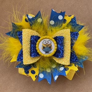 May include: A handmade hair bow featuring a Minion character. The bow is a combination of yellow, blue, and white ribbons, with a central image of a Minion. The bow is embellished with yellow feathers and glitter.