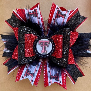 May include: A handmade hair bow featuring red, black, and white ribbons with a glittery finish. The bow includes a central emblem with a red and black logo, accented by black feathers. The bow is designed with a layered construction.