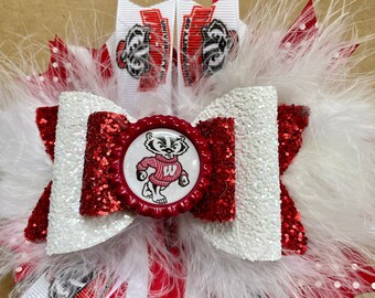 May include: A white and red glitter bow with a red and white striped ribbon and a white feather trim. The center of the bow features a red bottle cap with a white badger wearing a red sweater with a white "W" on it.