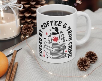 Op de afbeelding: Een witte keramische mok met een zwarte afbeelding van een stapel boeken met daarop een koffiekopje en een mes. De tekst luidt FUELED BY COFFEE & TRUE CRIME. Het ontwerp bevat rode bloedachtige spetters en kleine zwarte sterren.