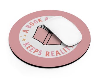Op de afbeelding: Een ronde, oudroze muismat met een witte cirkel in het midden, een roze boekillustratie en de tekst A BOOK KEEPS REALITY. Een witte computermuis ligt op het oppervlak van de mat en bedekt gedeeltelijk de afbeelding van het boek.