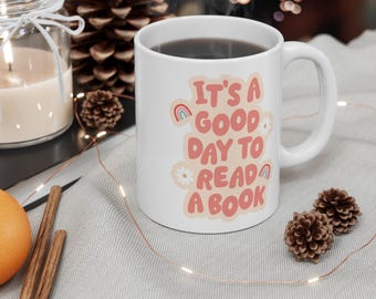 Op de afbeelding: Een witte keramische mok met de tekst "IT'S A GOOD DAY TO READ A BOOK" in roze letters, versierd met kleine illustraties van regenbogen en witte madeliefjes op een licht perzikkleurige achtergrondvorm.