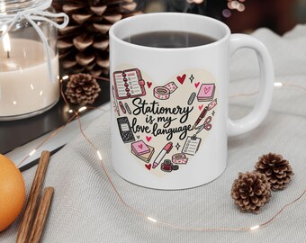 Op de afbeelding: Een witte keramische mok met een hartvormige afbeelding met diverse kantoorartikelen zoals notitieboekjes, pennen, tape en een schaar, met de zwarte tekst "Stationery is my love language" in het midden van het hart.