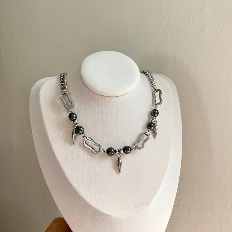 May include: A silver-toned necklace with a chain design, featuring dark gray beads and elongated, pointed charms. The necklace is displayed on a white bust, showcasing its design and details.