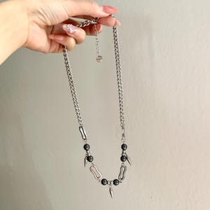 May include: Silver-toned chain necklace featuring black beads and decorative metal elements. The necklace has a curb chain design with a pendant of black beads and spike-shaped accents. The necklace is held up by a hand.