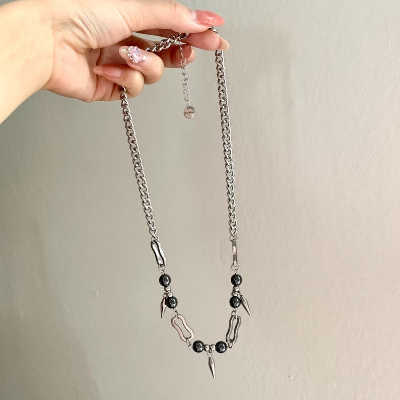 May include: Silver-toned chain necklace featuring black beads and decorative metal elements. The necklace has a curb chain design with a pendant of black beads and spike-shaped accents. The necklace is held up by a hand.