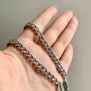 May include: A silver-colored chain necklace with a curb link design. The necklace is shown against a neutral background. The chain is made of a shiny, metallic material and has a secure clasp.
