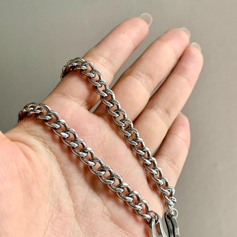 May include: A silver-colored chain necklace with a curb link design. The necklace is shown against a neutral background. The chain is made of a shiny, metallic material and has a secure clasp.