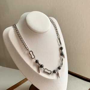 May include: A silver-toned necklace with a curb chain design. The necklace features dark gray beads and decorative metal elements, including elongated teardrop shapes and rectangular links. The jewelry is displayed on a white bust.
