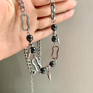 May include: A silver-toned necklace featuring a chain design with geometric links, dark gray beads, and pointed metal accents. The necklace is held up against a neutral background.