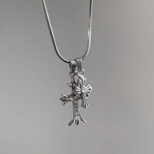 Puede incluir: Un collar de tono plateado con dos colgantes de cruz superpuestos en una cadena fina tipo serpiente. La cruz más grande está adornada con pequeñas piedras claras, mientras que la más pequeña presenta una piedra central y detalles ornamentados.