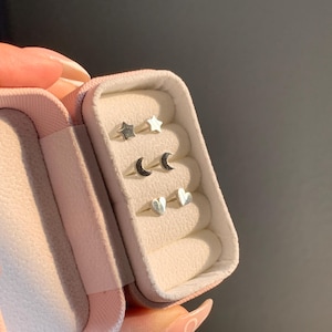 May include: A pink velvet jewelry box with three pairs of silver earrings. The earrings are shaped like stars, moons, and hearts.