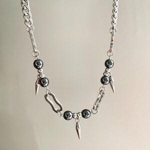 May include: A silver-toned necklace featuring a chunky chain design. The necklace incorporates dark gray, spherical beads and small, silver-colored spikes. The necklace has a modern, edgy aesthetic.