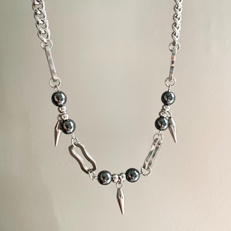 May include: A silver-toned necklace featuring a chunky chain design. The necklace incorporates dark gray, spherical beads and small, silver-colored spikes. The necklace has a modern, edgy aesthetic.
