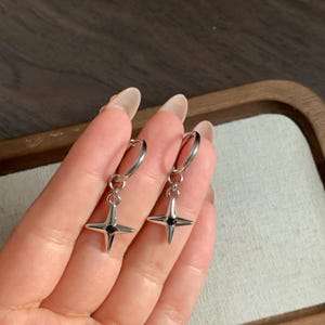 May include: A pair of silver hoop earrings with a four-pointed star pendant. Each star has a small, black center. The earrings are displayed against a neutral background, highlighting their minimalist design and metallic sheen.