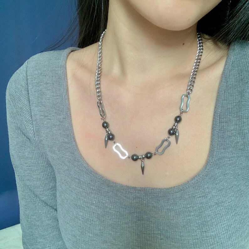 May include: Silver-toned chain necklace with a mix of geometric and bead elements. The necklace features a curb chain, oblong links, dark gray beads, and pointed cone-shaped pendants. The design is modern and edgy, suitable for various styles.