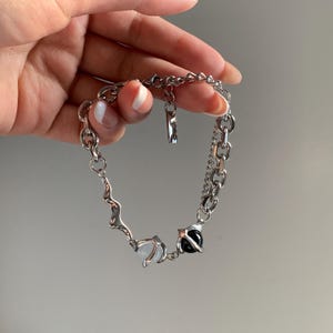May include: A silver-toned bracelet with a unique design. The bracelet features a combination of chain links and an abstract, wavy design. It includes a white and a black stone, adding contrast to the piece. The bracelet is held by a hand.