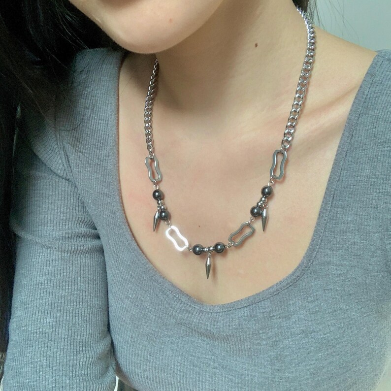 May include: A silver-toned necklace with a chain link design. The necklace features dark gray beads, elongated teardrop-shaped pendants, and open rectangular links. The necklace is worn against a gray shirt.