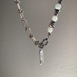 Può includere: Una collana a catena d'argento con un ciondolo d'argento e perline bianche. La catena ha un design a nodo e il ciondolo è un rettangolo lungo e sottile.