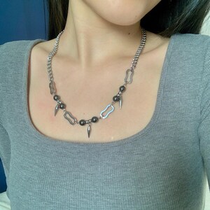 May include: Silver-toned necklace featuring a chunky chain and a pendant design. The necklace has a series of dark gray beads and silver-toned spikes. The necklace is worn on a person wearing a gray shirt.