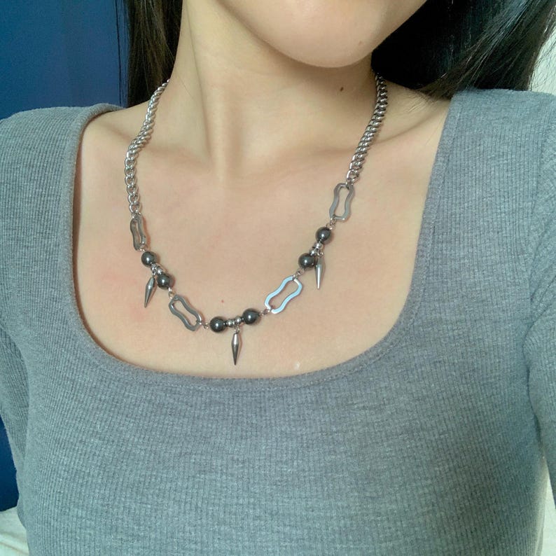 May include: Silver-toned necklace featuring a chunky chain and a pendant design. The necklace has a series of dark gray beads and silver-toned spikes. The necklace is worn on a person wearing a gray shirt.