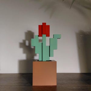 Può includere: Una scultura geometrica in legno di un cactus con un fiore rosso. Il cactus è verde chiaro e si trova in un vaso quadrato color rame. La scultura è su una superficie di legno scuro.