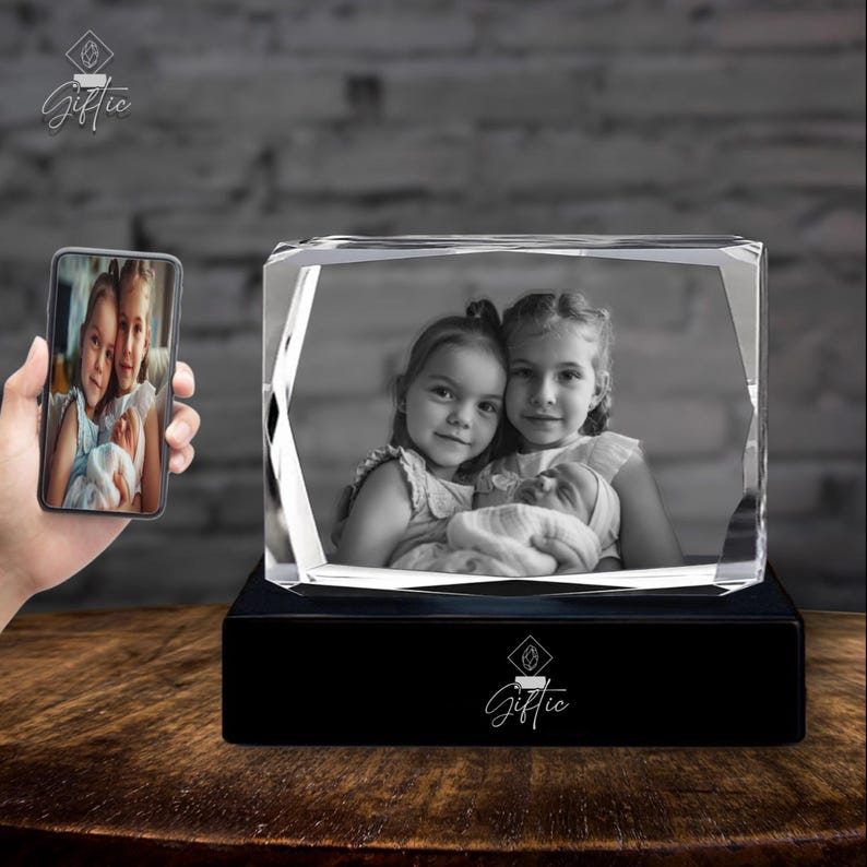 Op de afbeelding: Een 3D-kristallen kubus met een zwarte basis, met een zwart-witfoto van drie kinderen. De kristallen kubus wordt van onderen verlicht, waardoor een gloeiend effect ontstaat. De foto toont twee oudere zussen die een baby vasthouden.