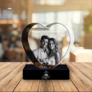Op de afbeelding: Een hartvormige kristallen fotolijst met een zwarte basis. De lijst toont een zwart-wit foto van een stel en de tekst "Happy Valentine's Day". De lijst staat op een houten oppervlak.